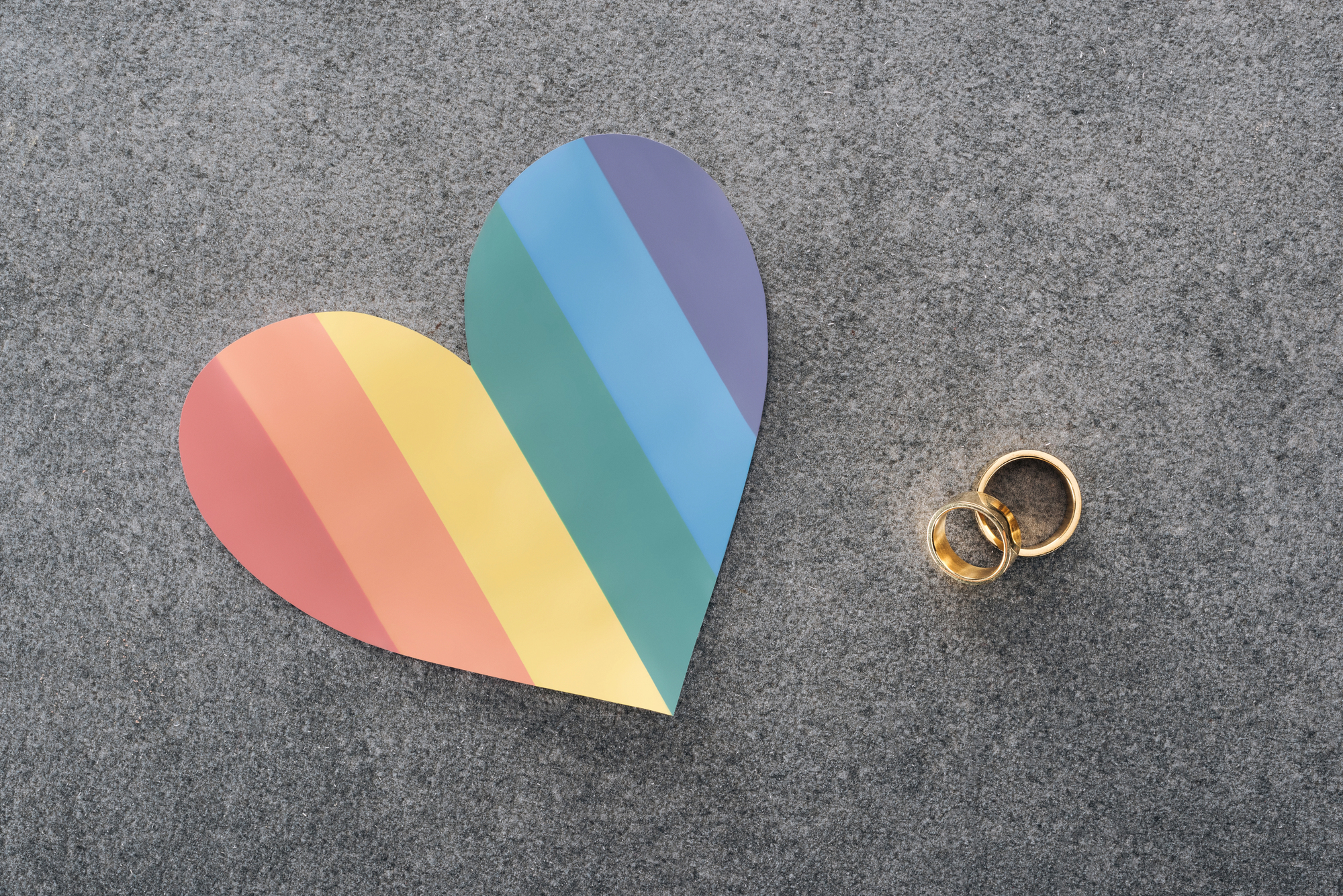 Pastel rainbow-striped heart on a gray carpet with gold wedding bands nearby.