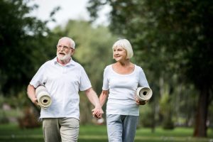 Older couple walking in a park, each holding a rolled yoga mat and wearing light clothing.