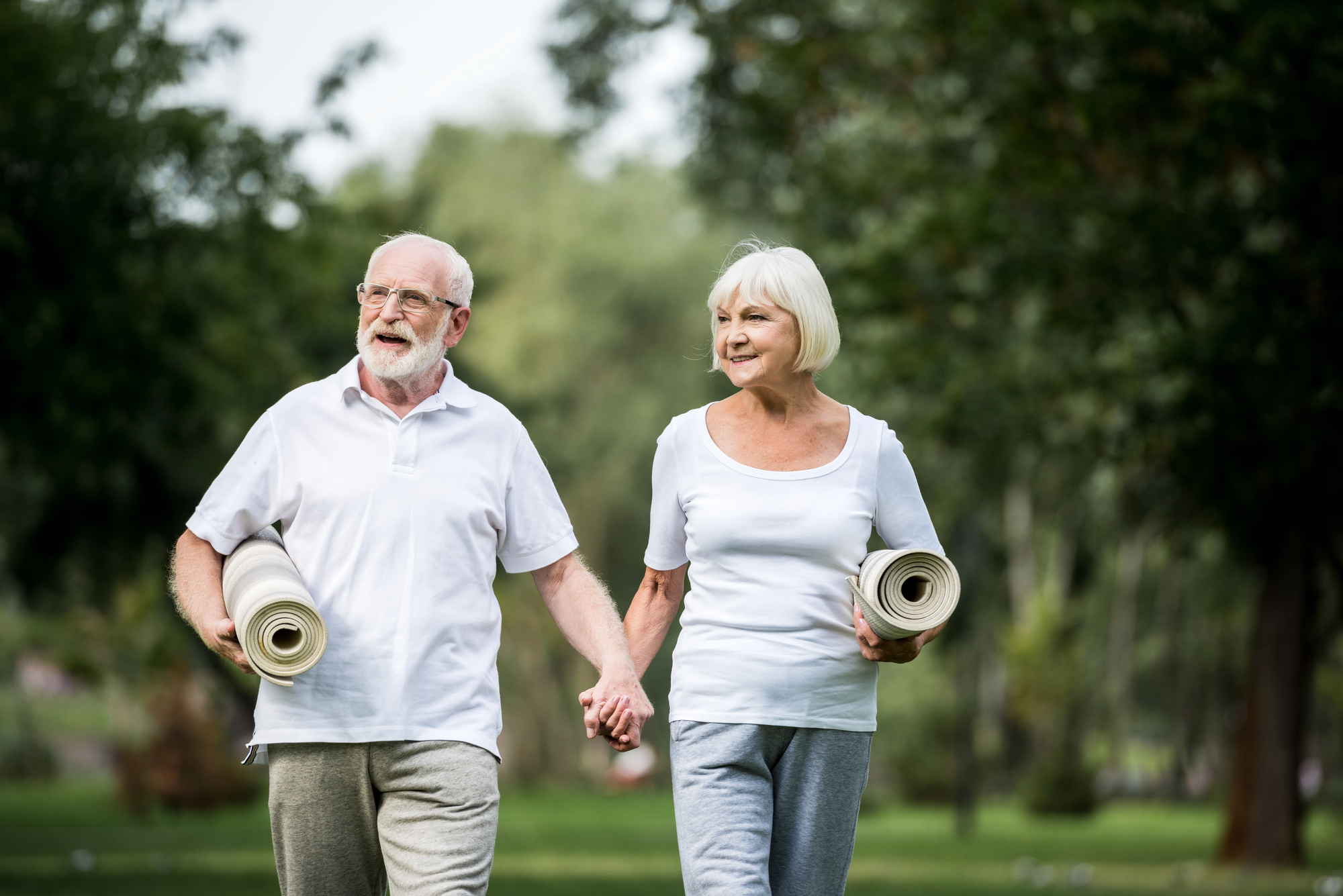 Older couple walking in a park, each holding a rolled yoga mat and wearing light clothing.