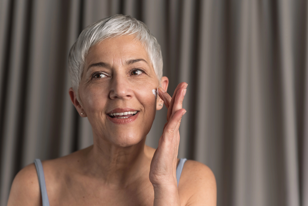 Older woman with short white hair applying moisturizer under her eye, smiling at the mirror.