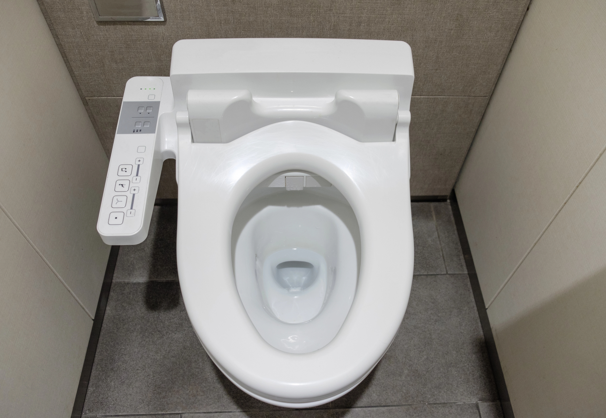 White elongated toilet with a side-mounted control panel (bidet features) visible on the left in a small bathroom stall, gray tiled floor and walls behind it.
