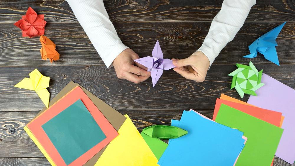 Hands fold a purple origami star on a dark wooden table, surrounded by colorful paper sheets and various origami shapes.