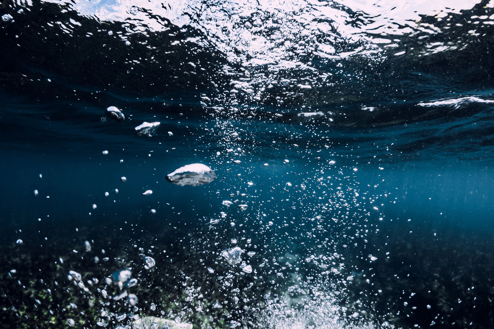 Underwater photo showing numerous bubbles rising from the bottom toward the sunlit surface in clear blue water