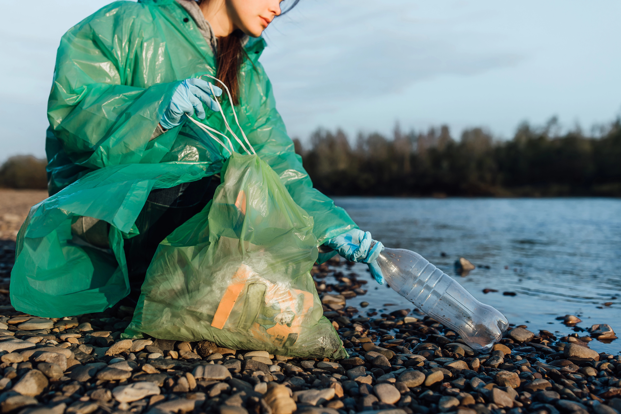 Person in a green plastic suit and blue gloves collects a plastic bottle on a rocky shoreline near the water.