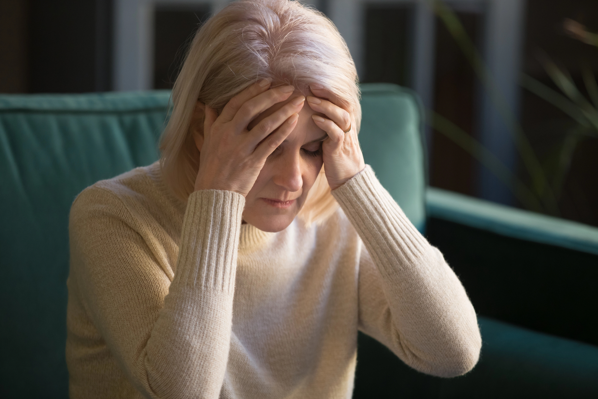 Woman with blond hair holds her head in distress, eyes closed, wearing a beige sweater in a teal chair setting