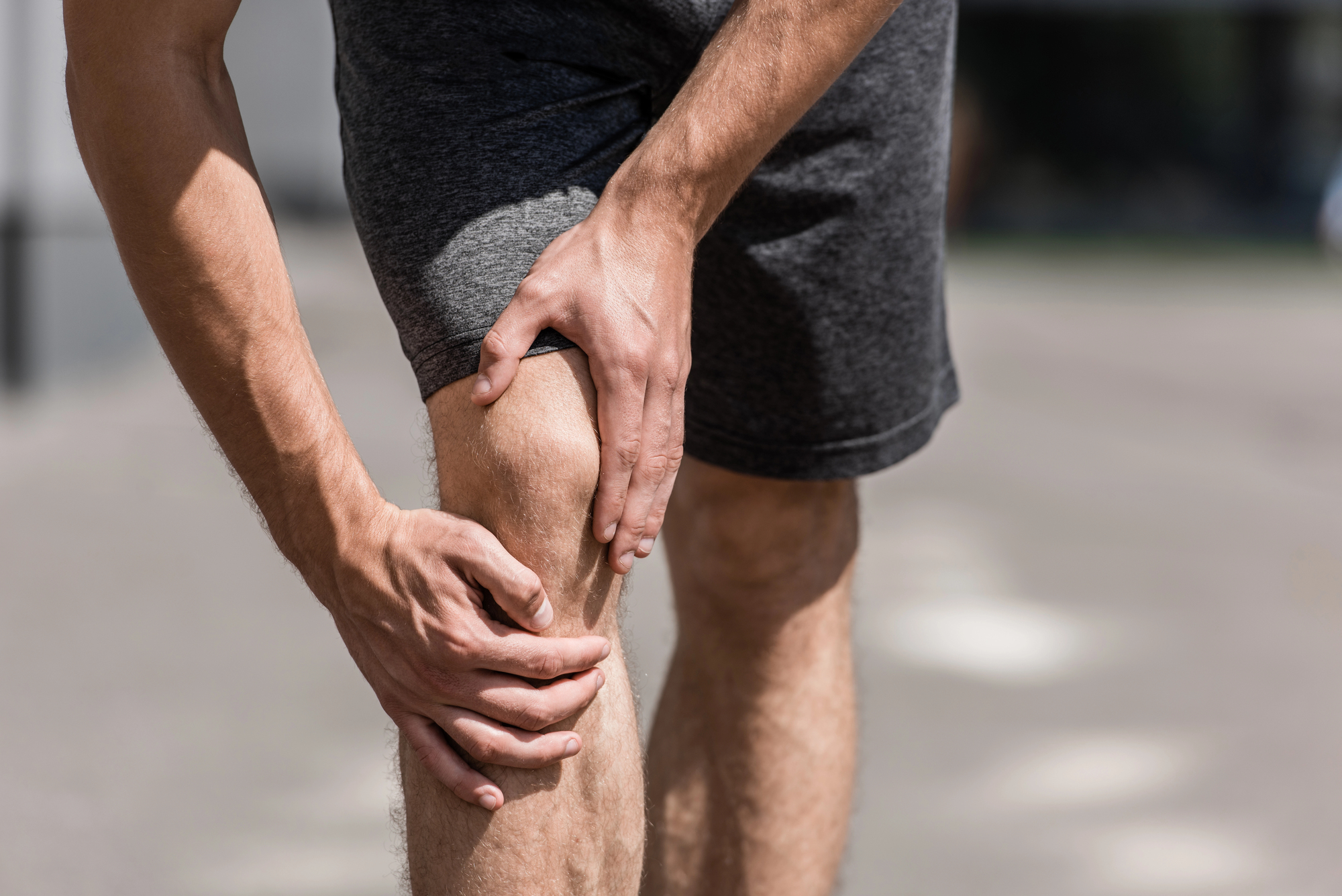 Close-up of a person in gray athletic shorts touching their knee, suggesting knee pain or discomfort.