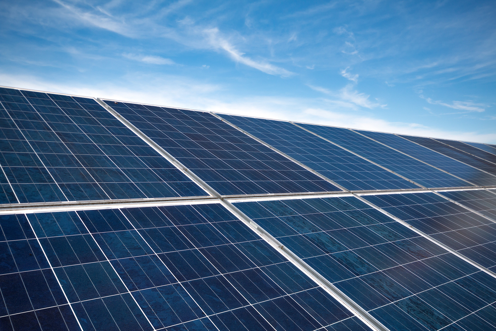 Rows of blue solar panels angled toward the sun under a clear blue sky on a sunny day, forming a solar farm grid.
