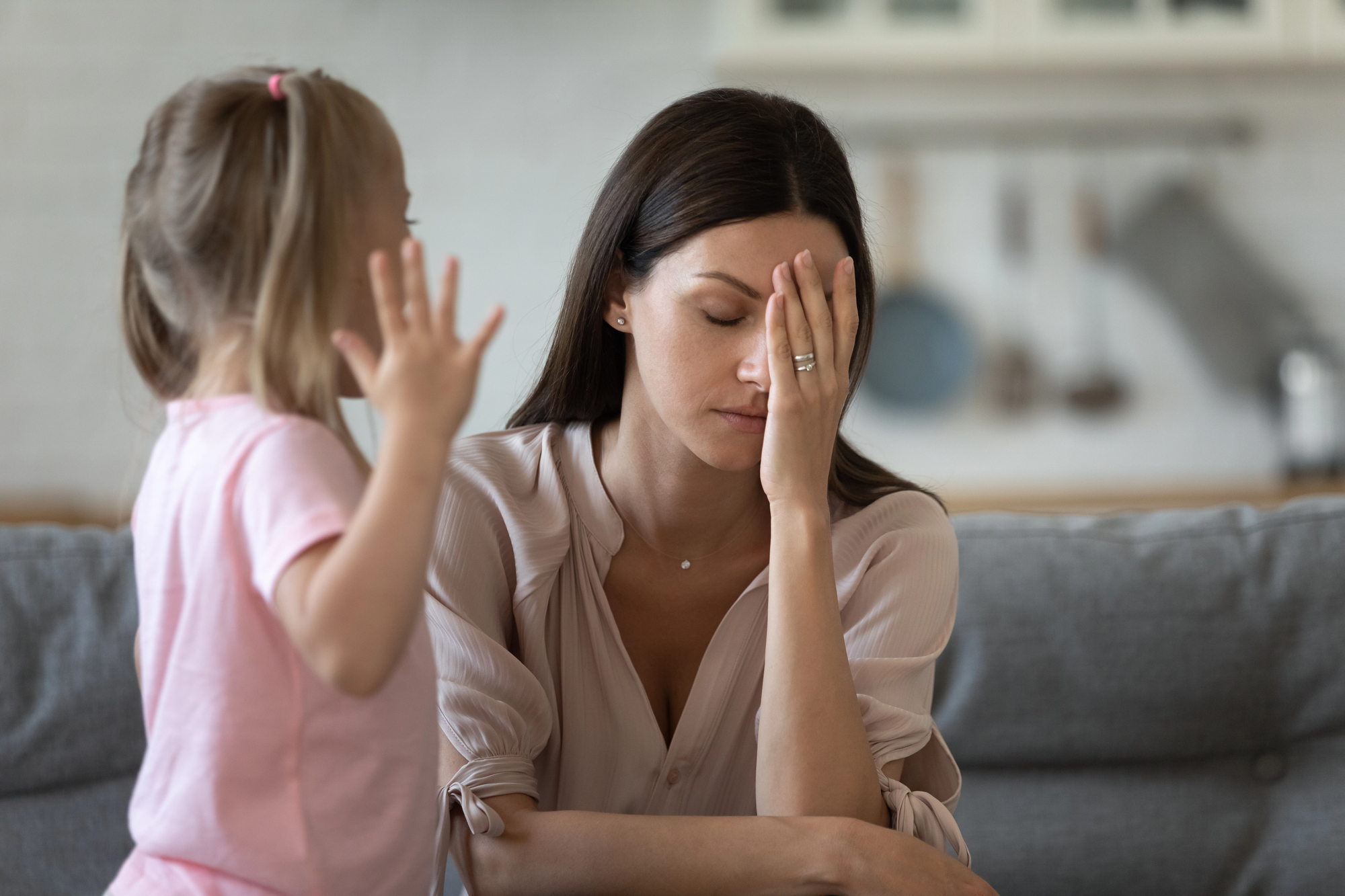 Woman sitting on a couch with her hand covering her face, looking stressed as a young girl reaches toward her in a pink shirt.