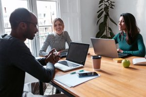 Three colleagues in a bright office have a meeting around a wooden table with laptops on it.