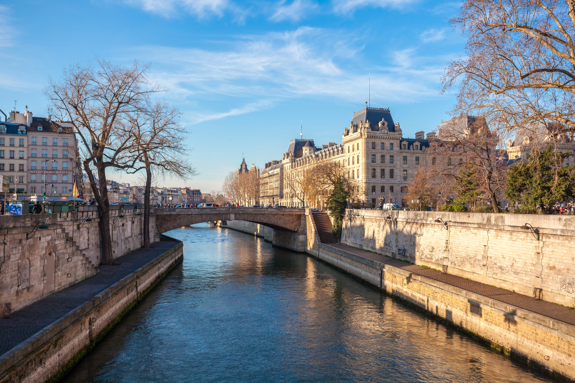 View of a sunlit Parisian riverfront with a stone embankment, ornate historic buildings, and a blue sky. Crowds are visible on the walkway and a bridge spans the river.