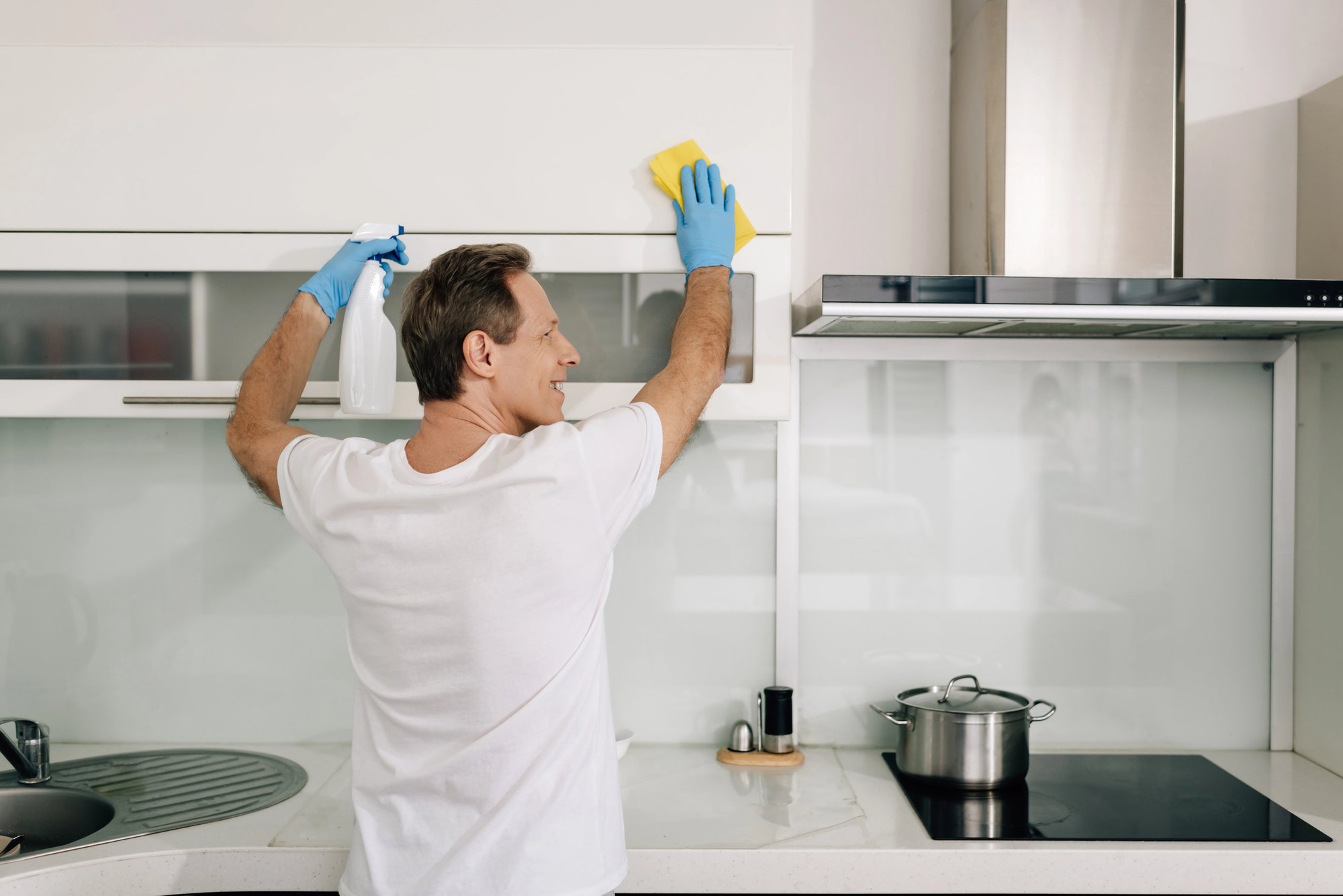 Man wearing a white T-shirt and blue gloves wipes a white kitchen cabinet with a yellow cloth while holding a spray bottle in the other hand.