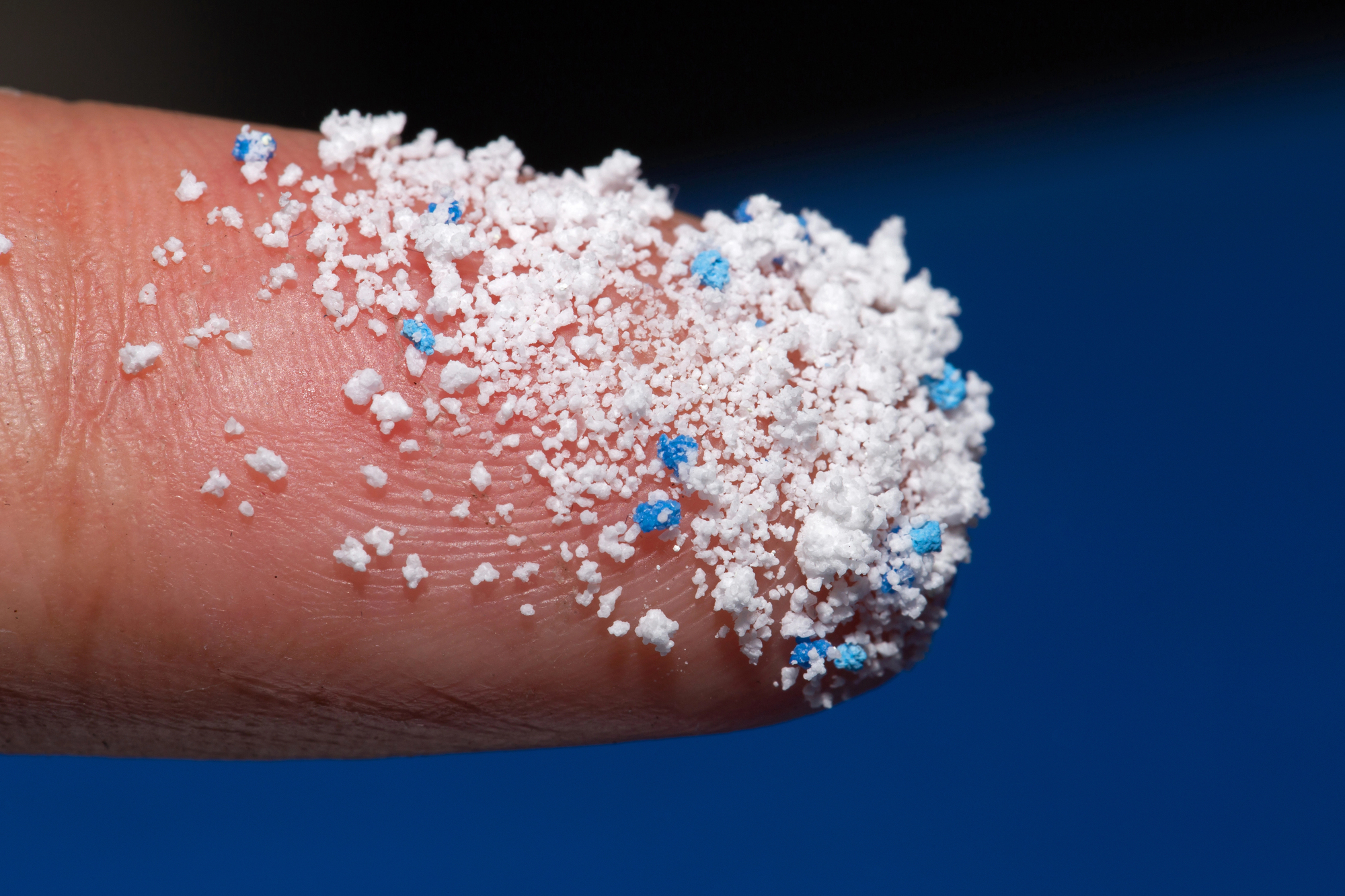 Close-up of a finger covered in white bath salt granules with blue specks, against a blue background