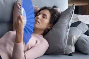 Woman lying on a gray couch with eyes closed, fanning herself with a blue handheld fan for cooling.