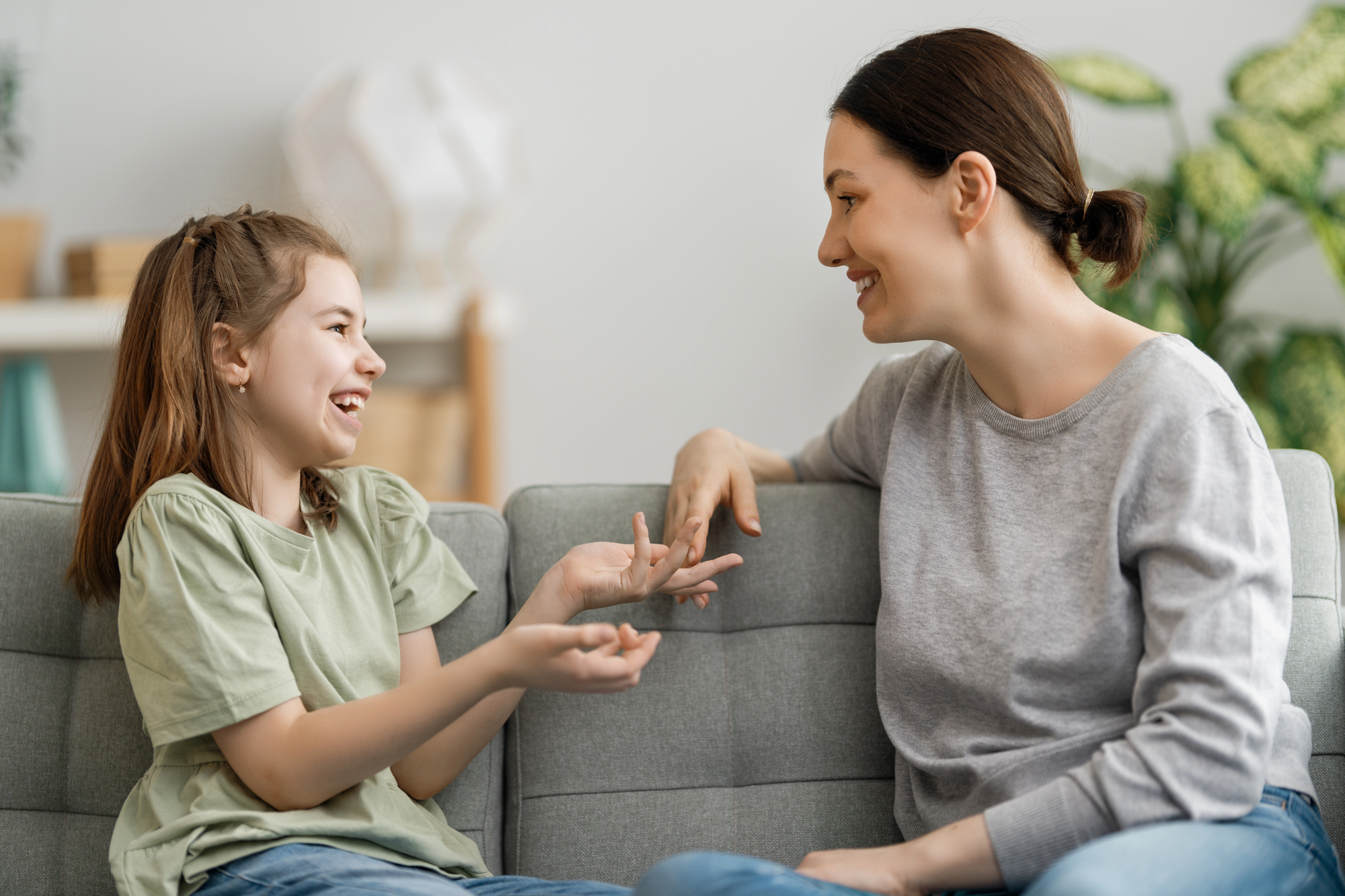 Mother and daughter sit on a gray sofa, smiling and playing with their fingers together at home.