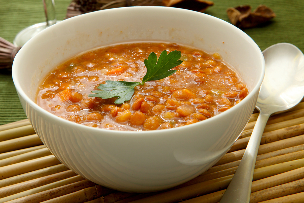 White bowl of lentil soup with carrots, garnished with a parsley leaf, on a wooden placemat with a spoon nearby.
