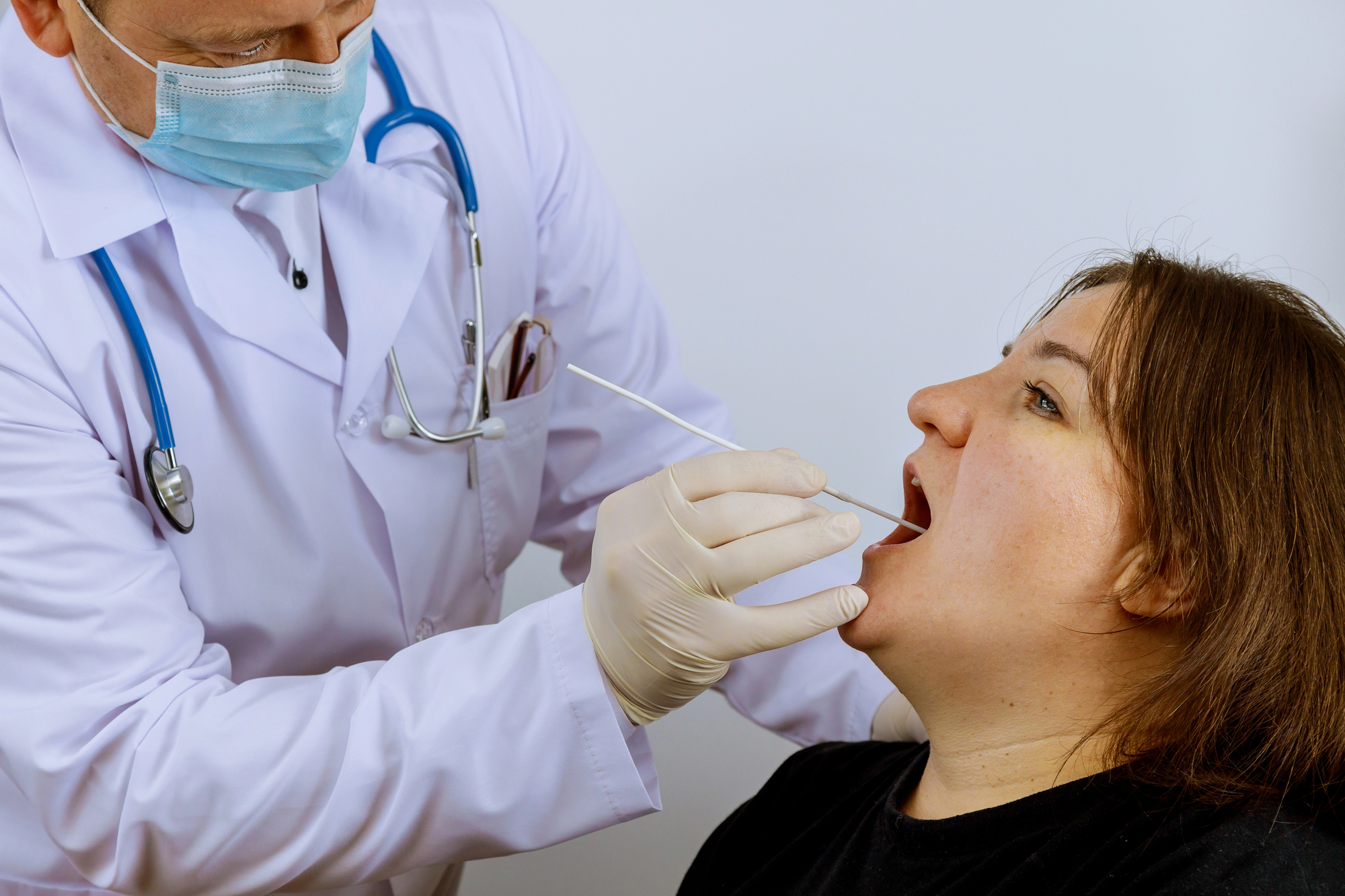 Medical professional in a mask swabbing a patient's throat with a long swab during a checkup, patient seated.