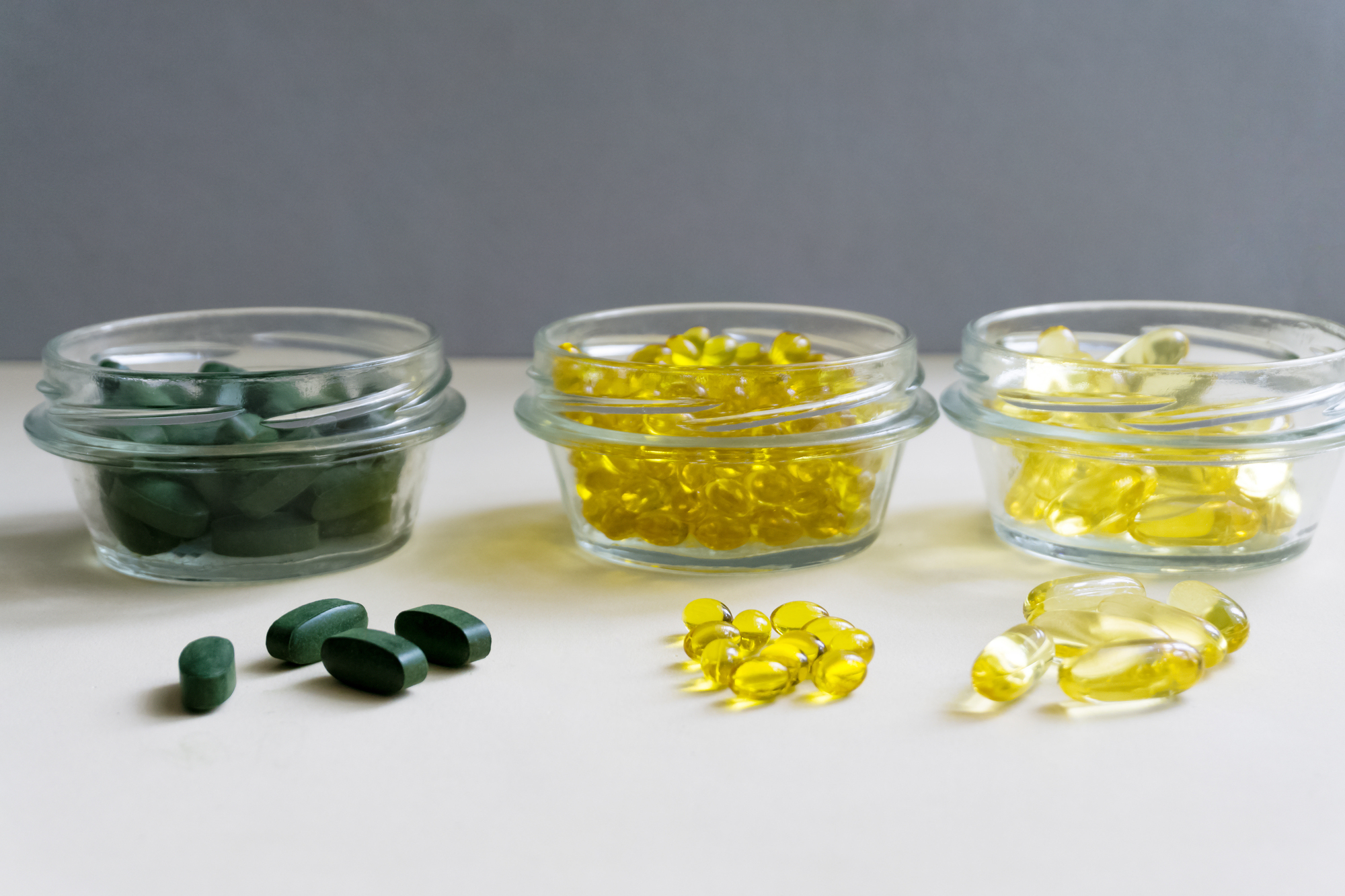 Three glass bowls holding different pills: dark green tablets, yellow gelatin capsules, and pale yellow soft capsules on a white surface.