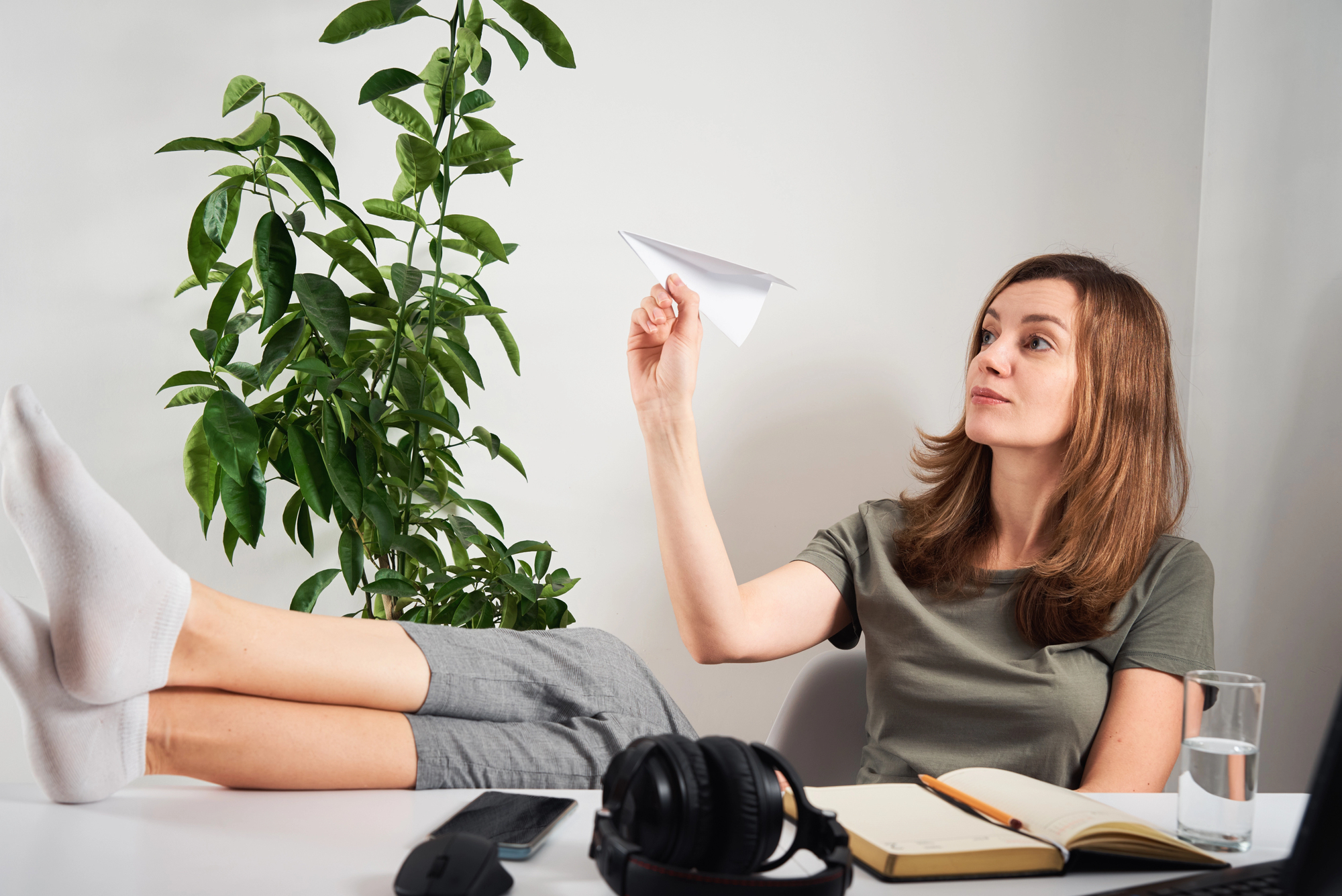 Woman sits at a desk with a paper plane in the air, plant in the background, and office items on the table.