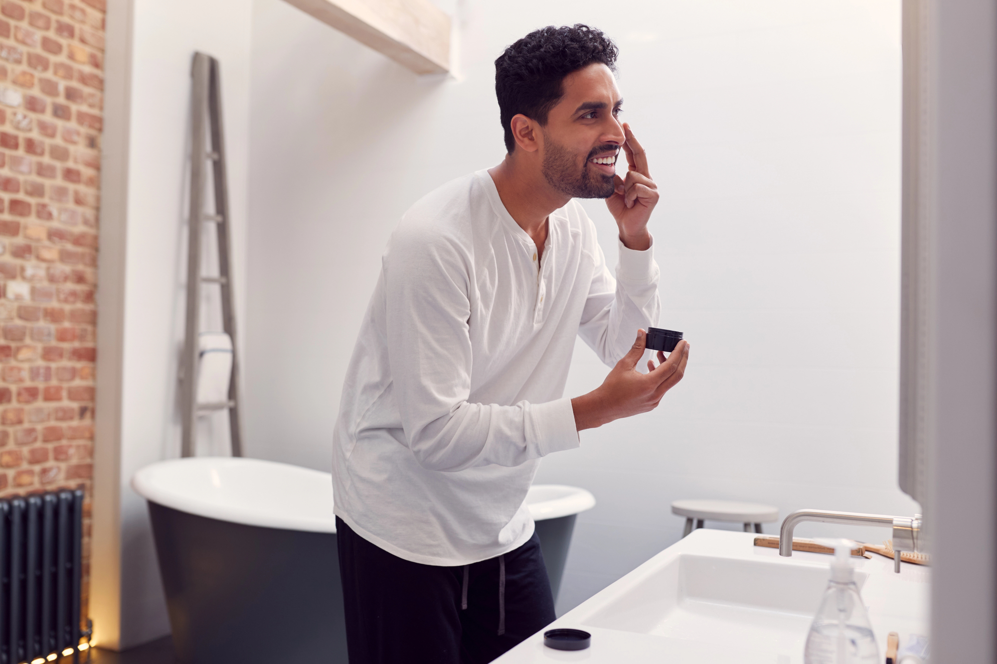 Man in a white long-sleeve shirt applies facial cream from a jar near his eye in a bright bathroom with a tub and brick wall on the left