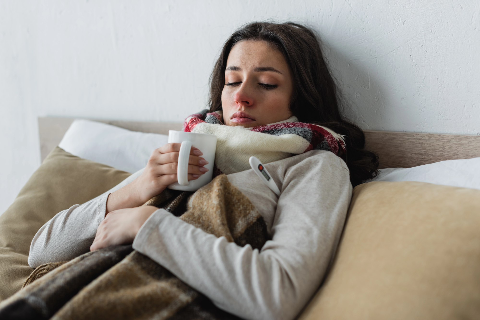 Woman resting in bed wrapped in a blanket, holding a mug and looking unwell.