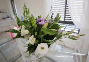 Bouquet of white roses with purple chrysanthemums and greenery wrapped in white netting on a glass table in a bright room with blinds in the background.