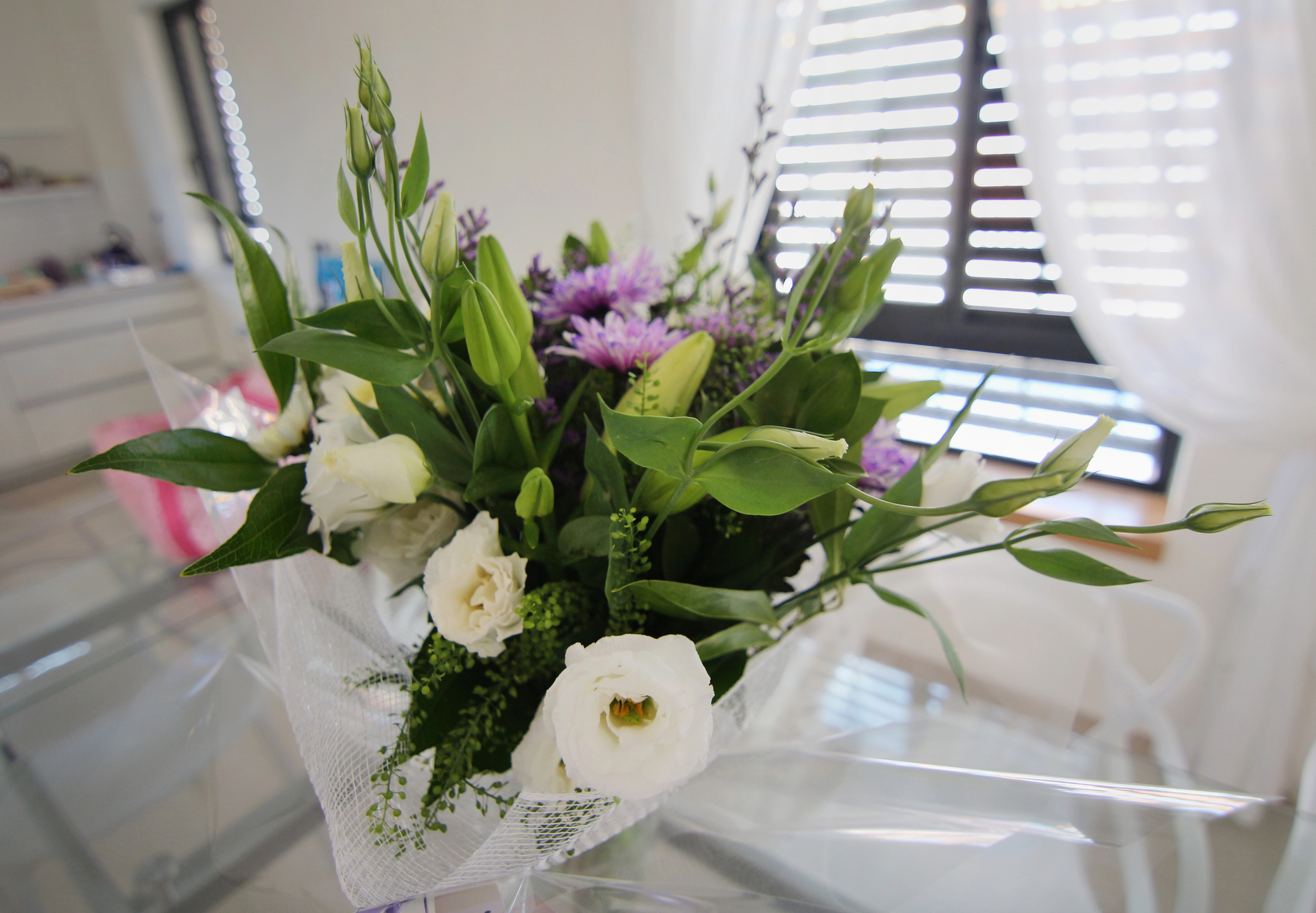 Bouquet of white roses with purple chrysanthemums and greenery wrapped in white netting on a glass table in a bright room with blinds in the background.