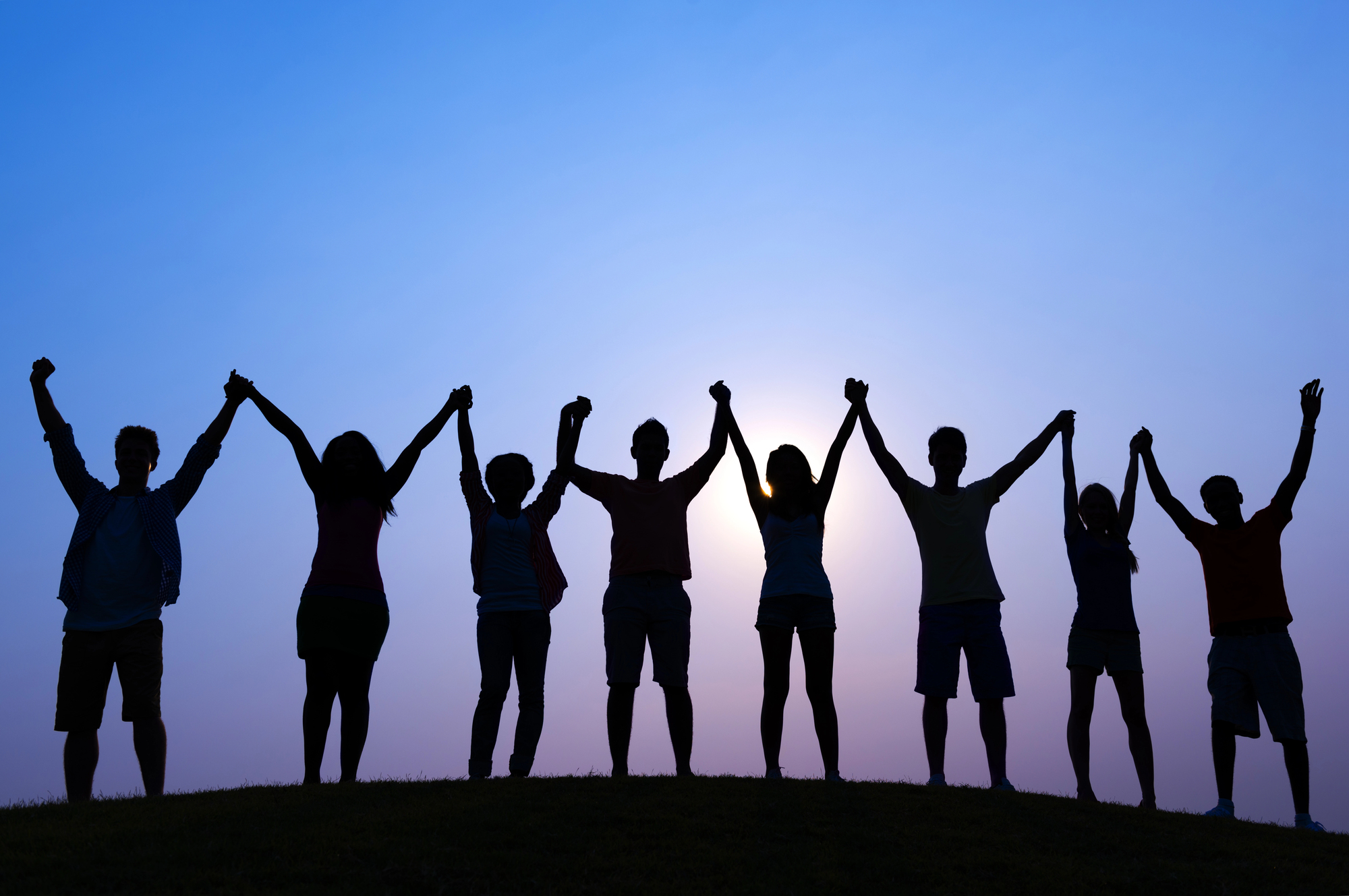 Silhouetted group of people holding hands overhead in a line on a hill at sunset.