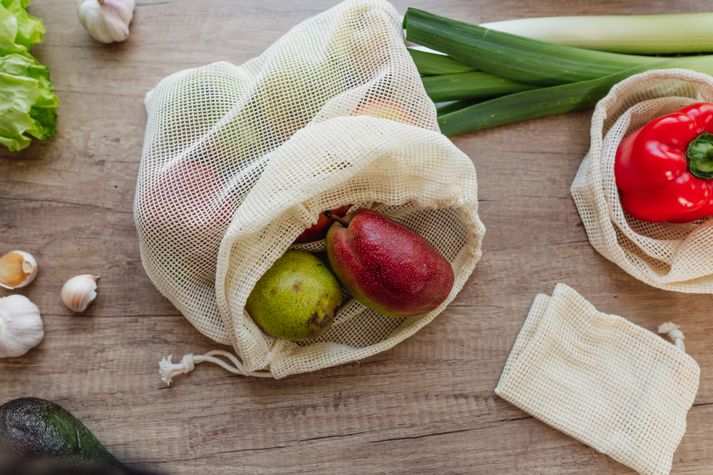 Reusable mesh produce bag opened on a wooden table with pears inside, surrounded by veggies (leeks, red pepper, garlic, lettuce) and another bag nearby.
