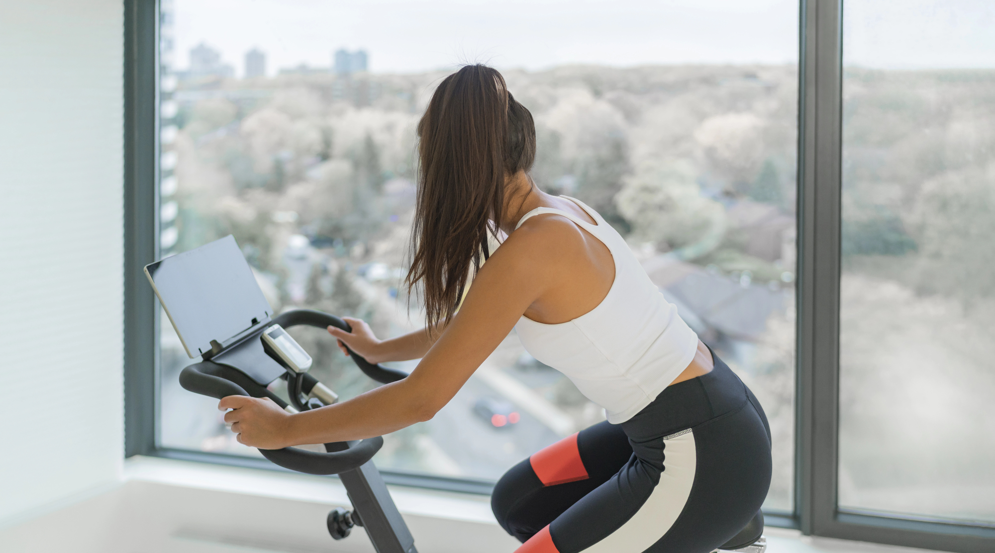 Woman wearing a white athletic top rides a stationary bike indoors beside large windows looking out at a cityscape.