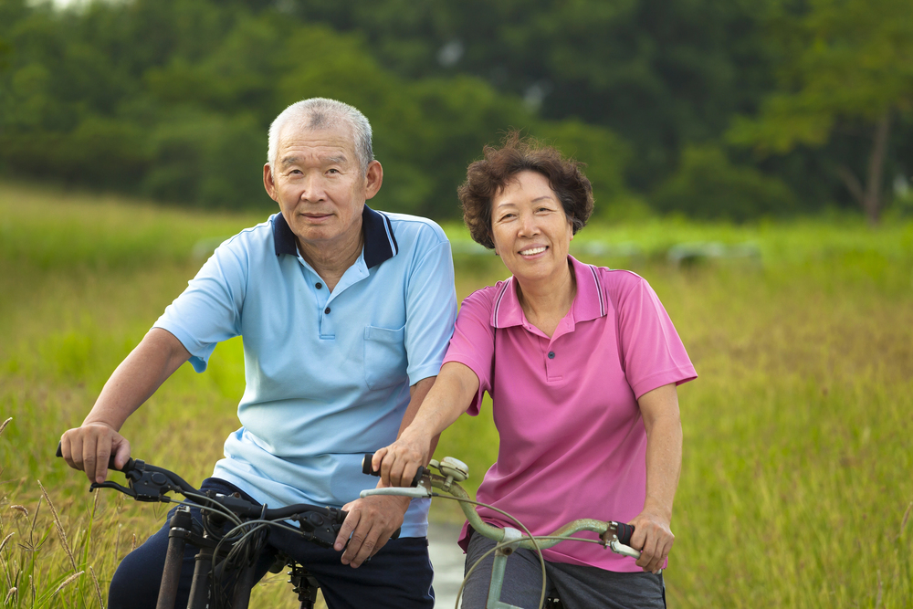 Older couple riding bicycles in a grassy countryside field, smiling at the camera.