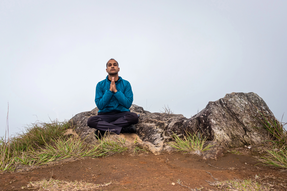 Person meditating on a rocky outcrop, seated cross‑legged with hands pressed together in prayer, wearing a blue jacket against a misty sky.