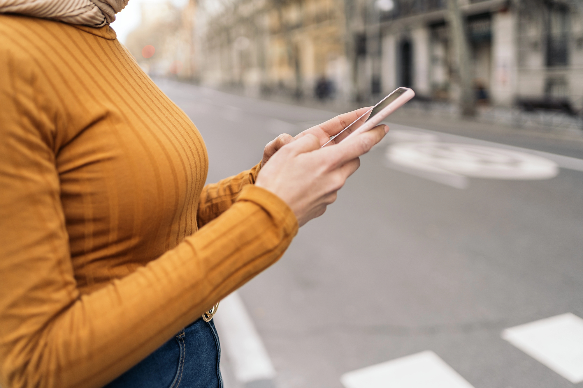 Person wearing an orange striped sweater holds a pink smartphone outdoors on a city street, hands in foreground.