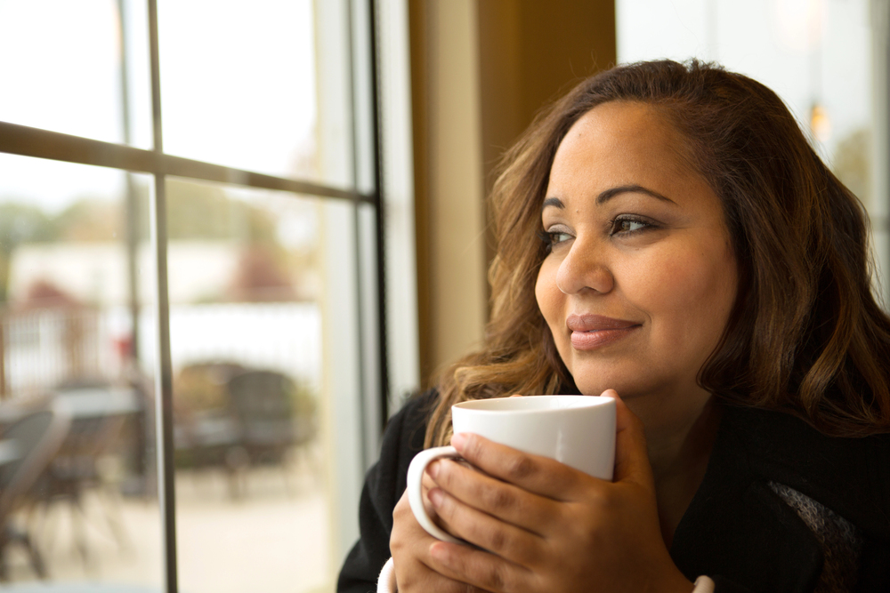 Woman sits by a window, holding a white mug and gazing thoughtfully outside in warm light.