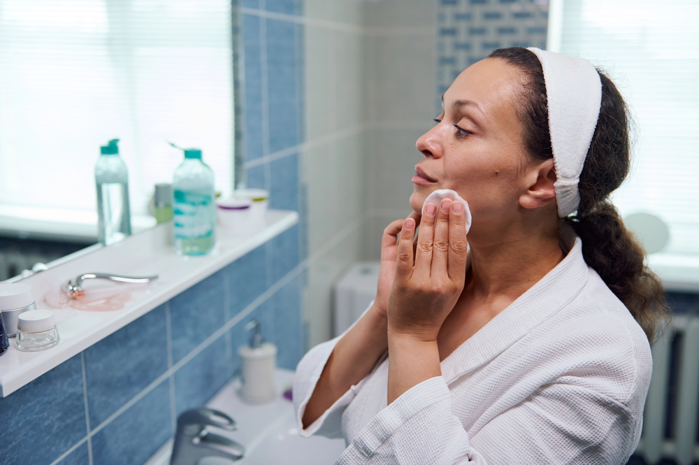 Woman in a white robe applies skincare with a cotton pad in a bathroom mirror area. Organized shelves with bottles in the background.