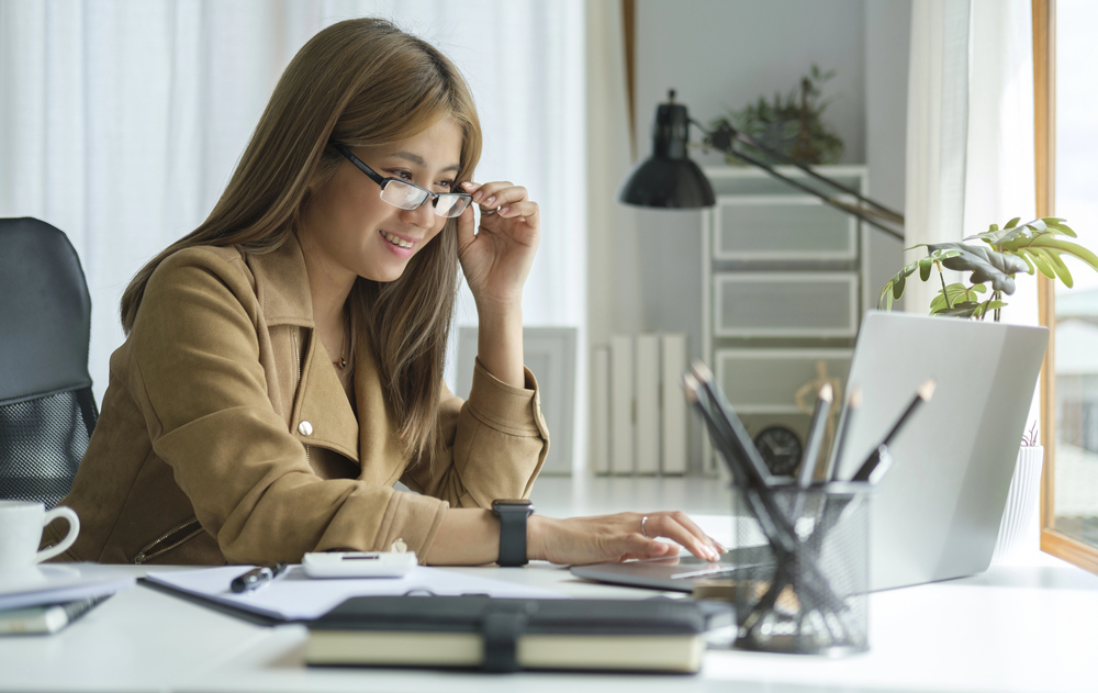 Woman wearing glasses works at a white desk with a laptop, notebook, and pencils nearby in a bright office.
