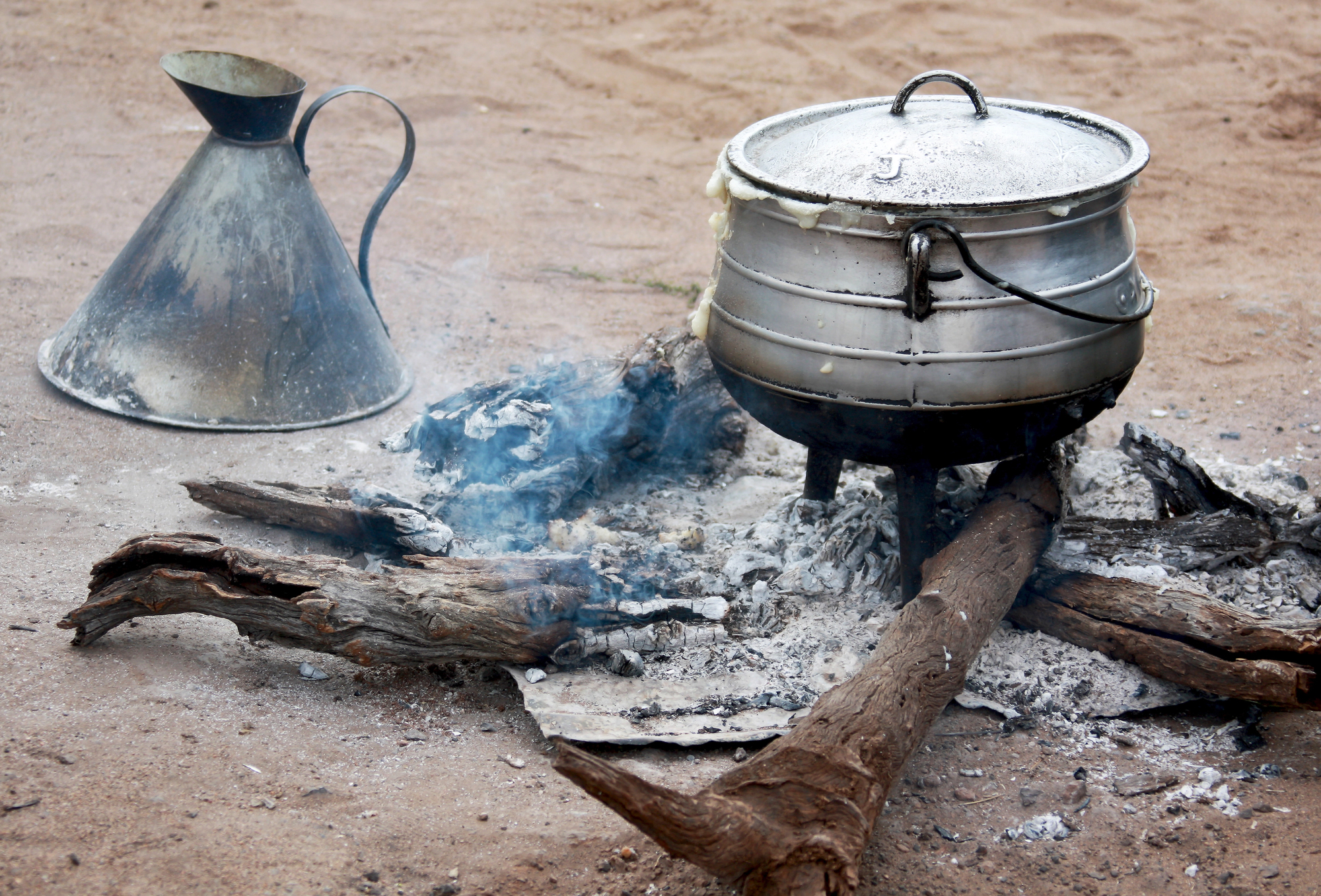 Old metal kettle beside a stacked steam pot on a campfire in a sandy outdoor area.