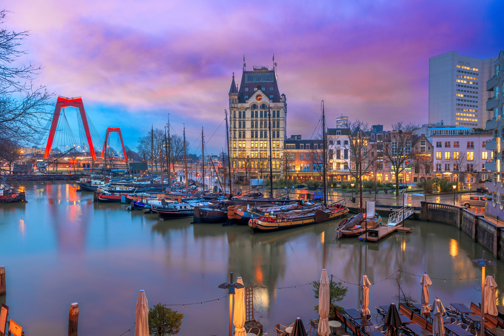Harbor scene at dusk with moored sailboats, a bright red arch bridge, and a historic riverside building.