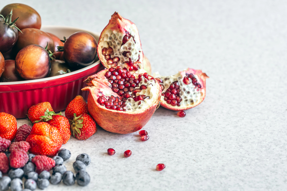 Fresh fruit display with pomegranate halves, a bowl of dark purple tomatoes, and a mix of strawberries, raspberries, and blueberries on a gray surface.