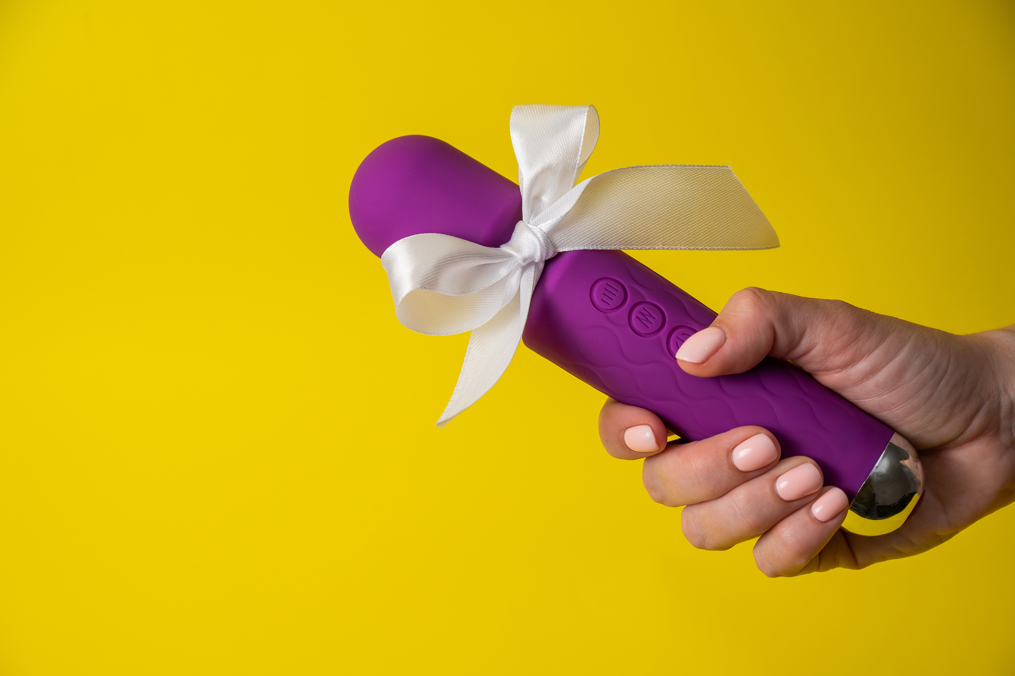 Purple vibrator held in a hand with a white satin bow against a bright yellow background (gift-like presentation).