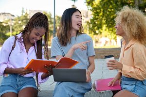 Three friends sit on outdoor steps, smiling and chatting with a notebook, a laptop, and a coffee cup nearby.
