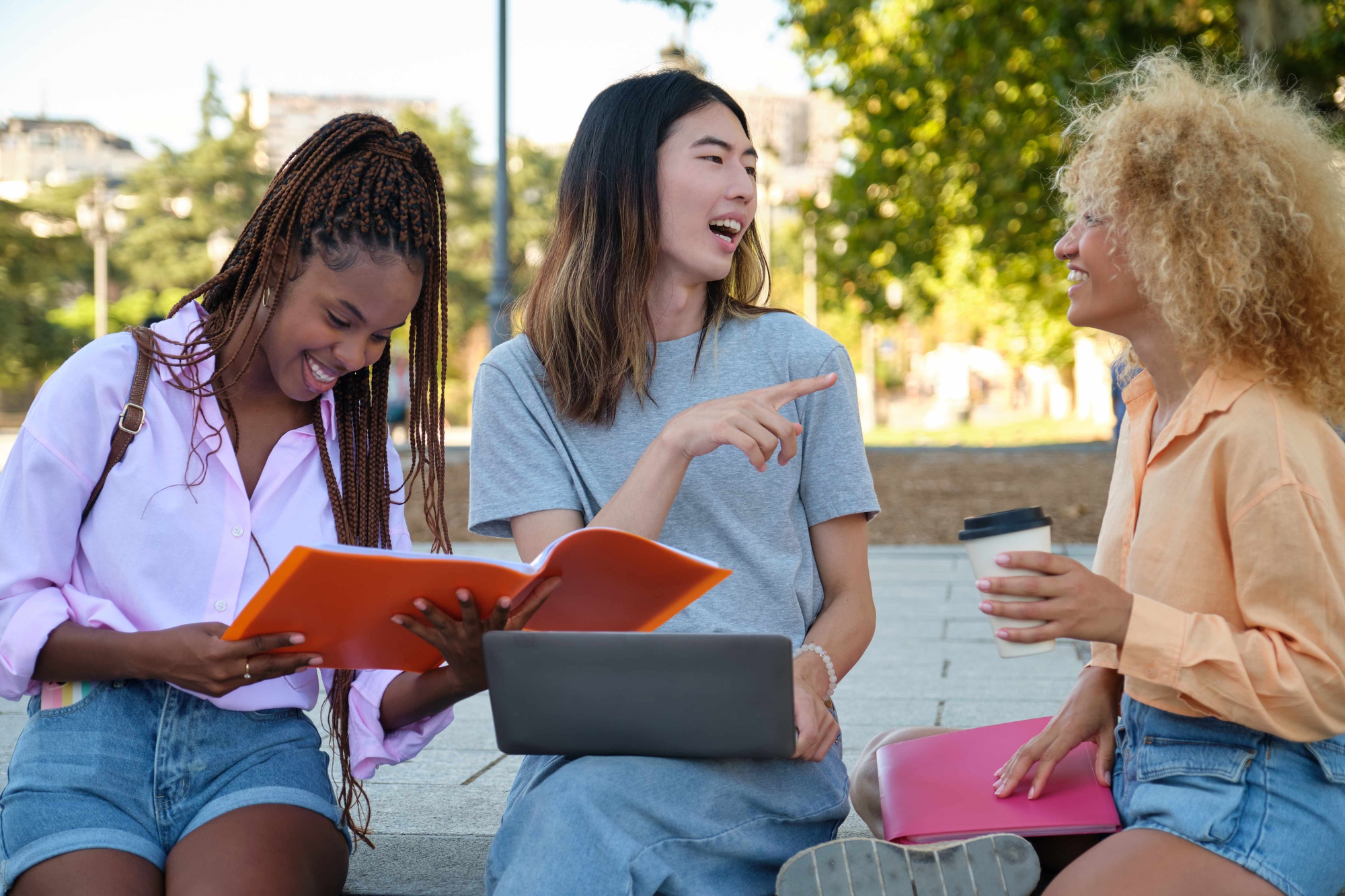 Three friends sit on outdoor steps, smiling and chatting with a notebook, a laptop, and a coffee cup nearby.