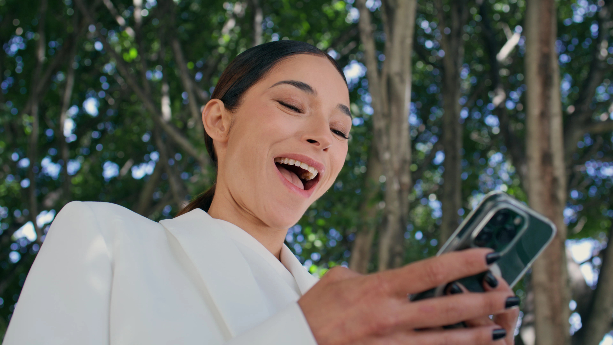 Woman outdoors, smiling while looking at her smartphone screen in a selfie pose.