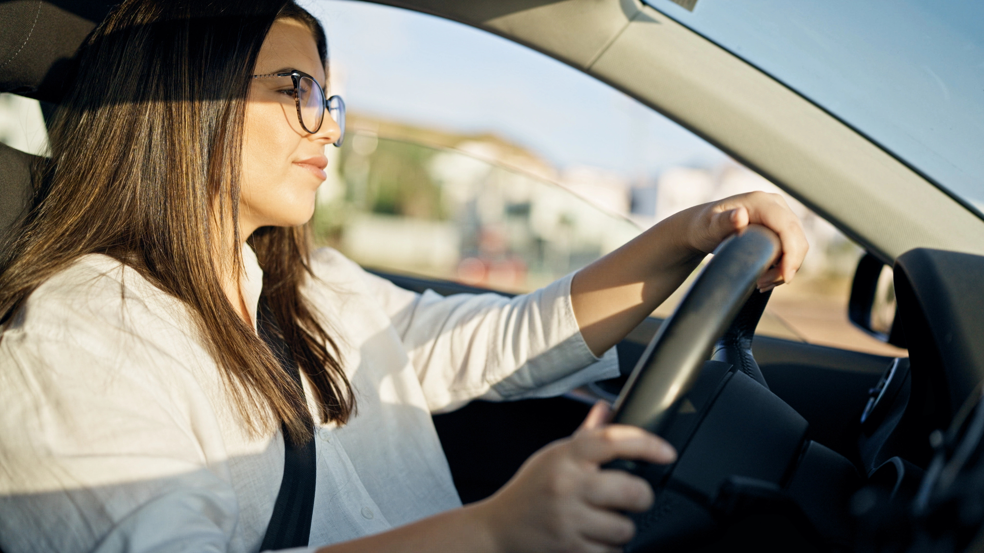 Woman with long hair and glasses driving a car, hands on the steering wheel, sunlight on her face.