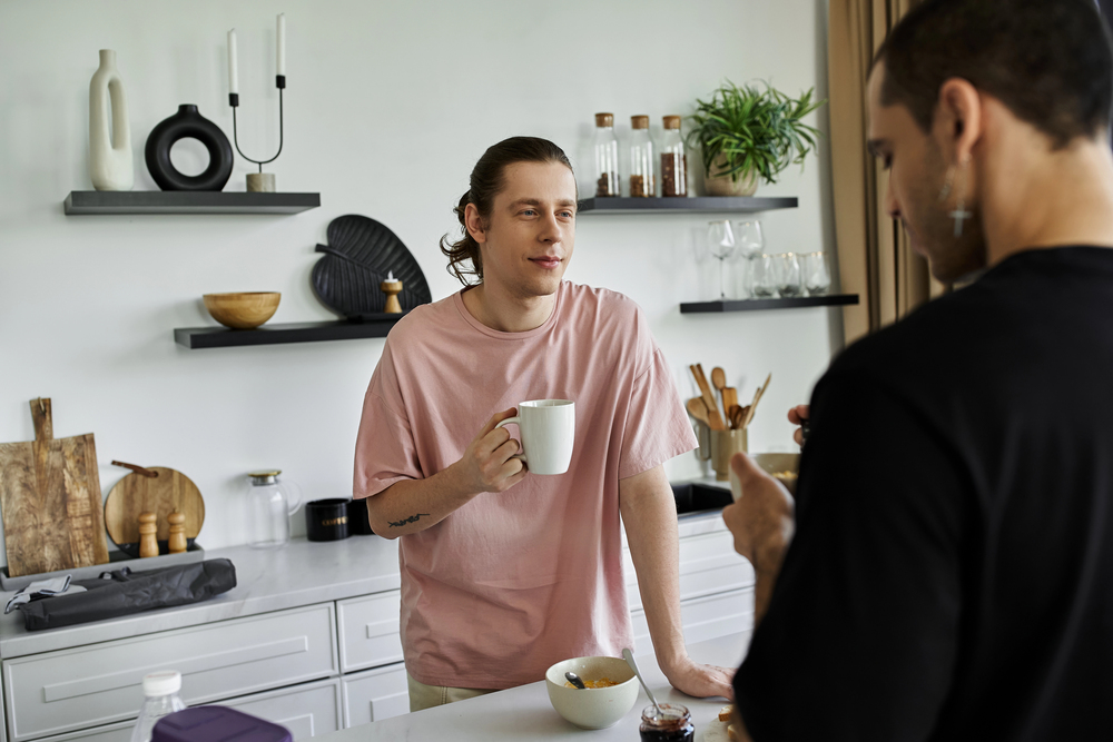Two men chat in a bright modern kitchen; one holds a mug while the other, facing away, gestures with a spoon near a bowl on the counter.