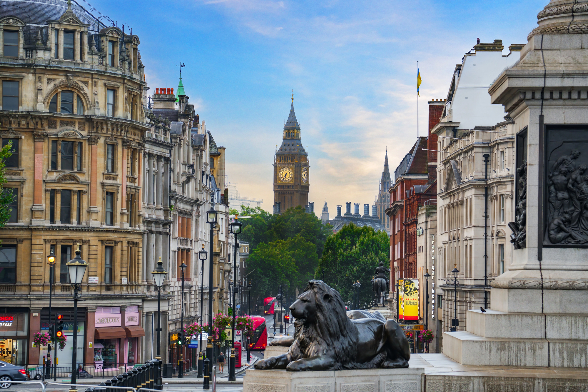 London street lined with historic façades, with Big Ben in the distance and stone lions at the foreground monument.
