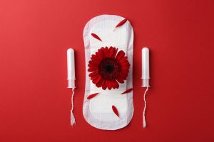 White sanitary pad laid on a red background, decorated with a bright red gerbera flower and scattered red petals for a feminine themed image.
