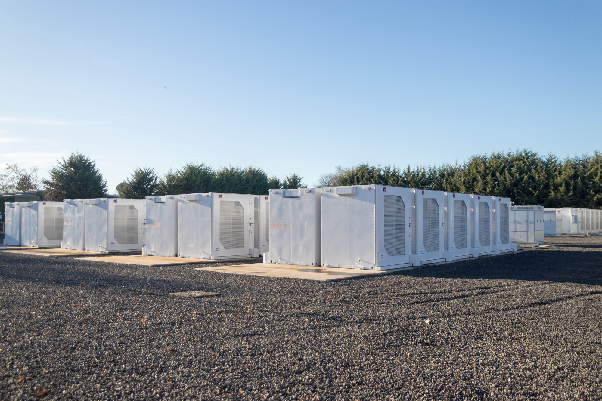 Row of white modular industrial containers with ventilation panels on a gravel lot, trees in the background against a clear blue sky.
