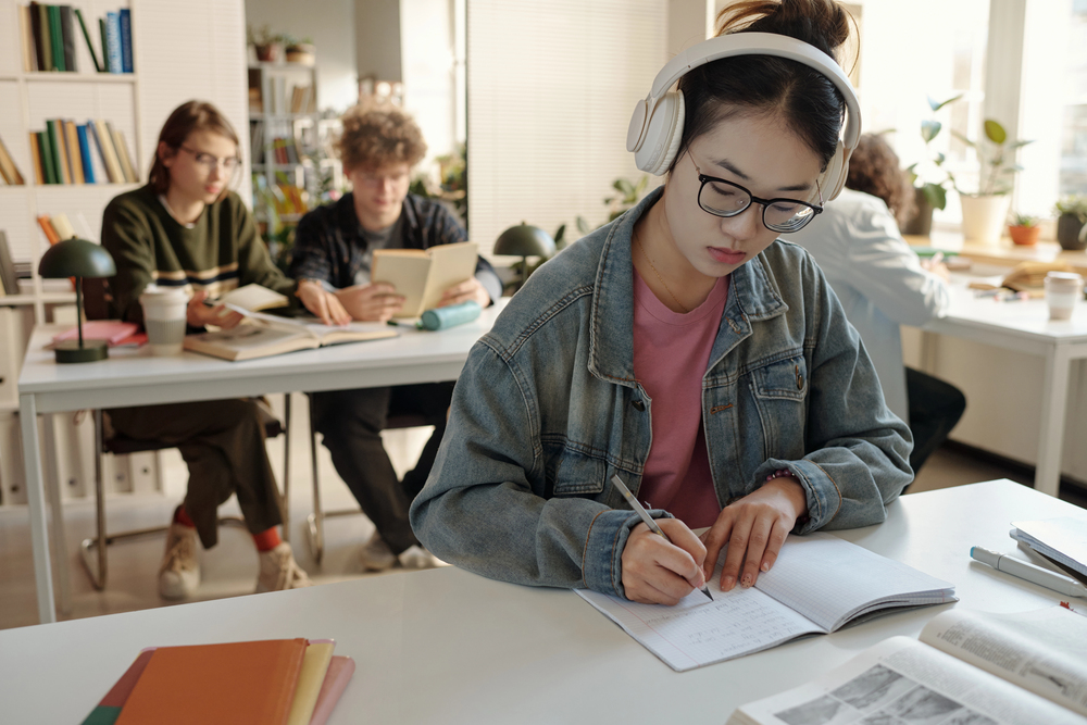 Student with headphones and glasses writes in a notebook at a desk while others read in a bright library.