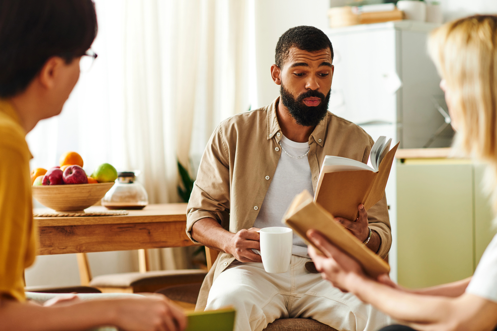 Bearded man in a beige shirt reads a book while holding a mug, with two friends nearby in a bright kitchen setting.