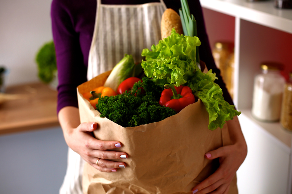 Person in an apron holds a paper bag overflowing with fresh vegetables (lettuce, kale, peppers) and bread in a kitchen.